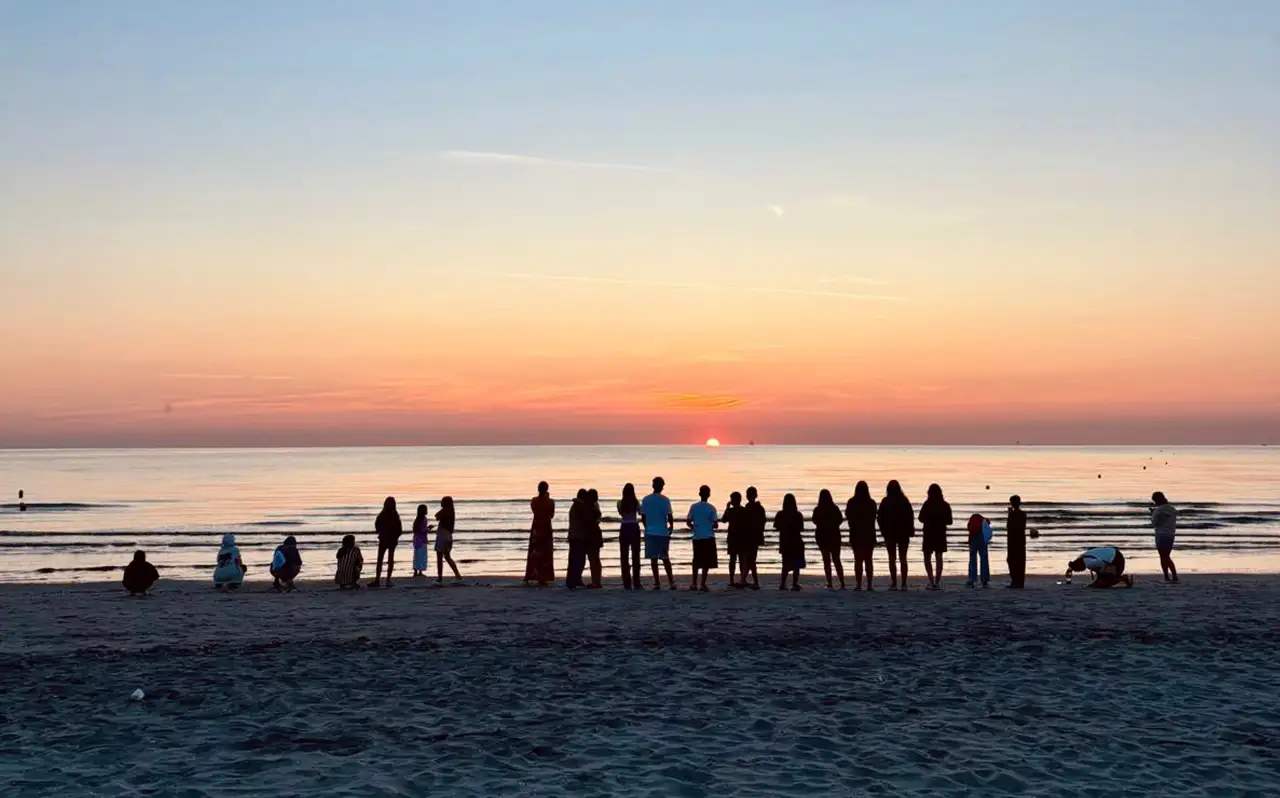 Zdjęcie przedstawiające uczestników obozu językowego we Włoszech, ustawionych w rzędzie na plaży, na tle morza i wschodzącego słońca